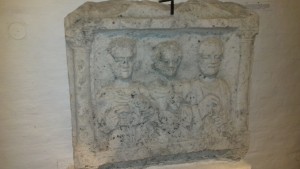 Mészkő sírsztélé. Kétoldalt korinthosi féloszlopokkal szegélyezett mezőben három férfialak, előttük középen egy gyermek mellképe. A bal oldali alak derekán katonai öv (cingulum - Kr. u. 3. század).